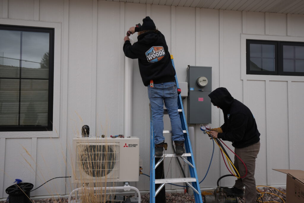 Woods Heating and Cooling technicians repairing a Mitsubishi Electric ductless mini-split, checking refrigerant pressure with gauges.