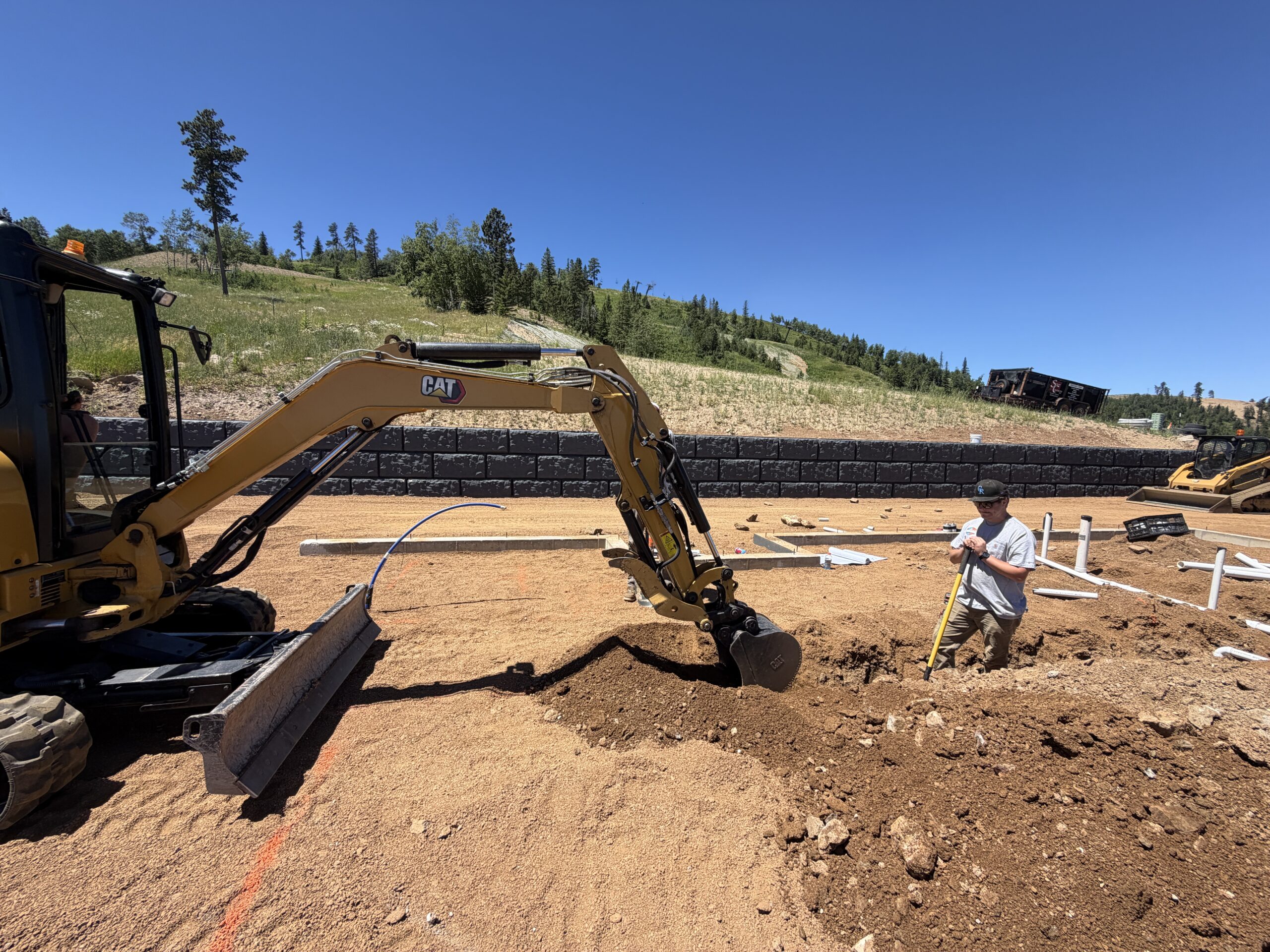 Mini excavator digging a trench beside a man with a shovel at a new construction site near Deadwood, SD, with PVC pipe stubbed up from the foundation.