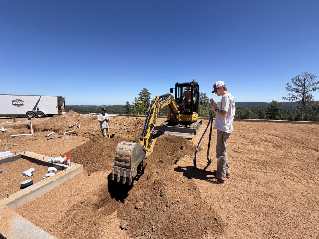 Woods Heating and Cooling plumbing crew staging PVC drain fittings and trenching for underground rough-in beside the company trailer on a Black Hills new build.
