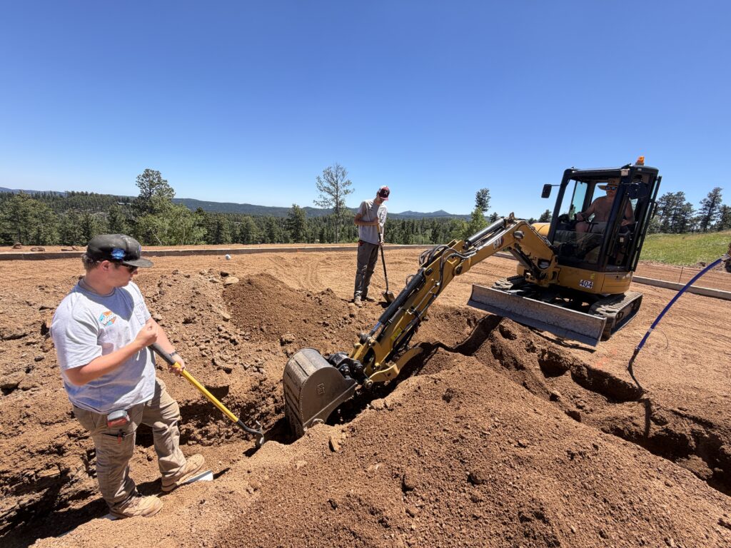 Woods Heating and Cooling crew and a mini excavator trenching through rocky soil for plumbing rough-in on a mountain lot near Lead, SD.